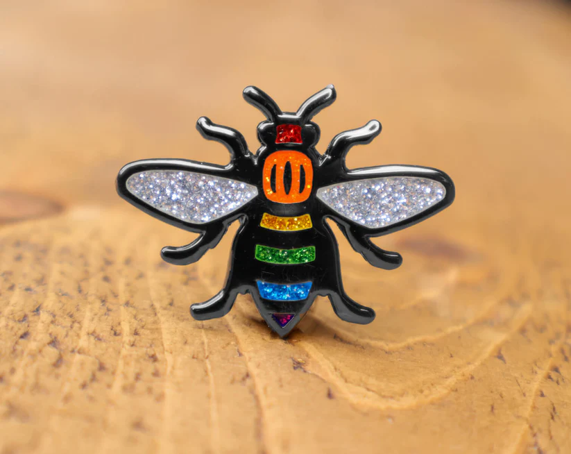 A rainbow glitter bee enamel pin on a wooden table.