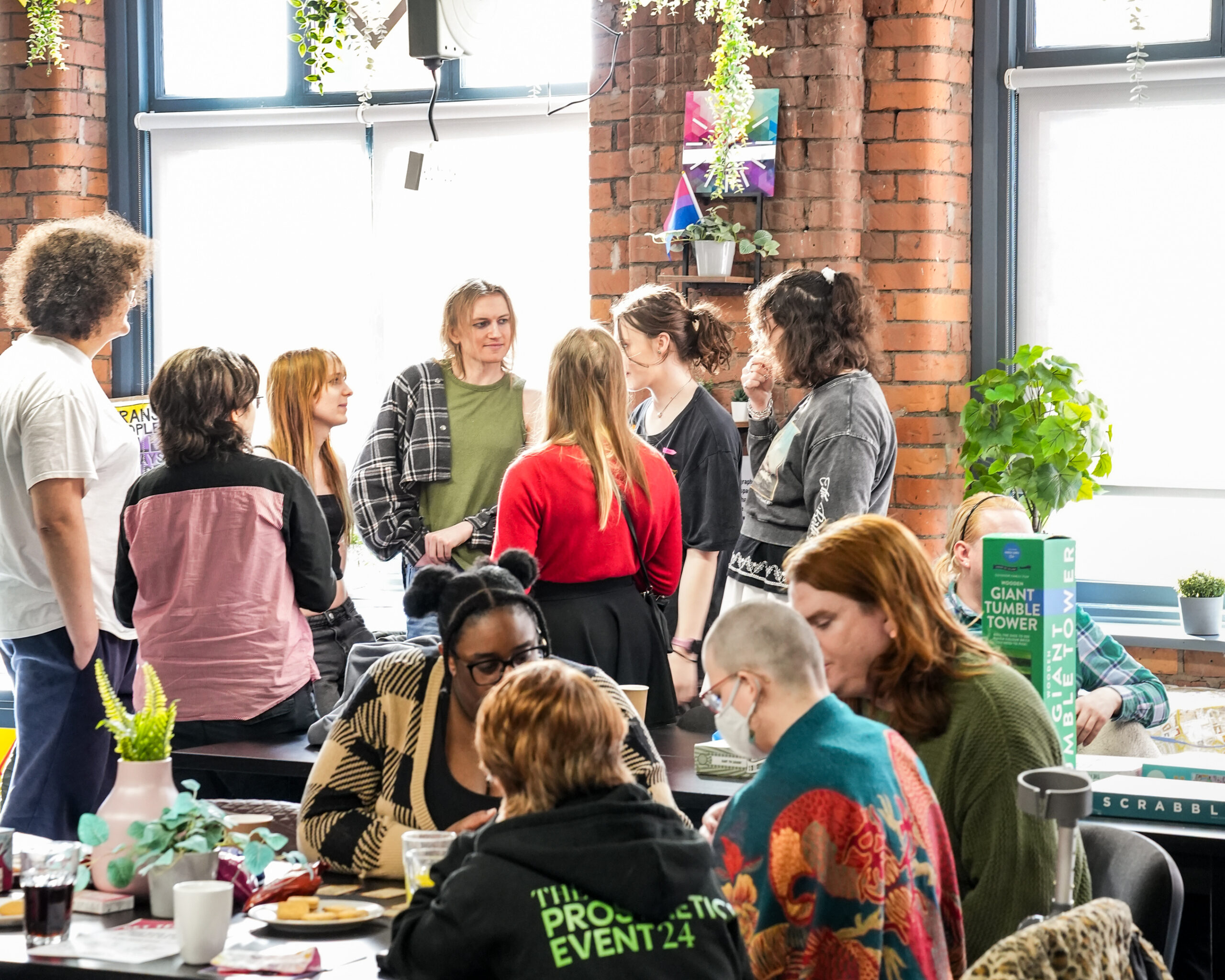 A group of LGBTQ+ people standing and chatting in a circle in LGB Foundation's centre. In the lower foreground, another group is sitting and doing an activity.