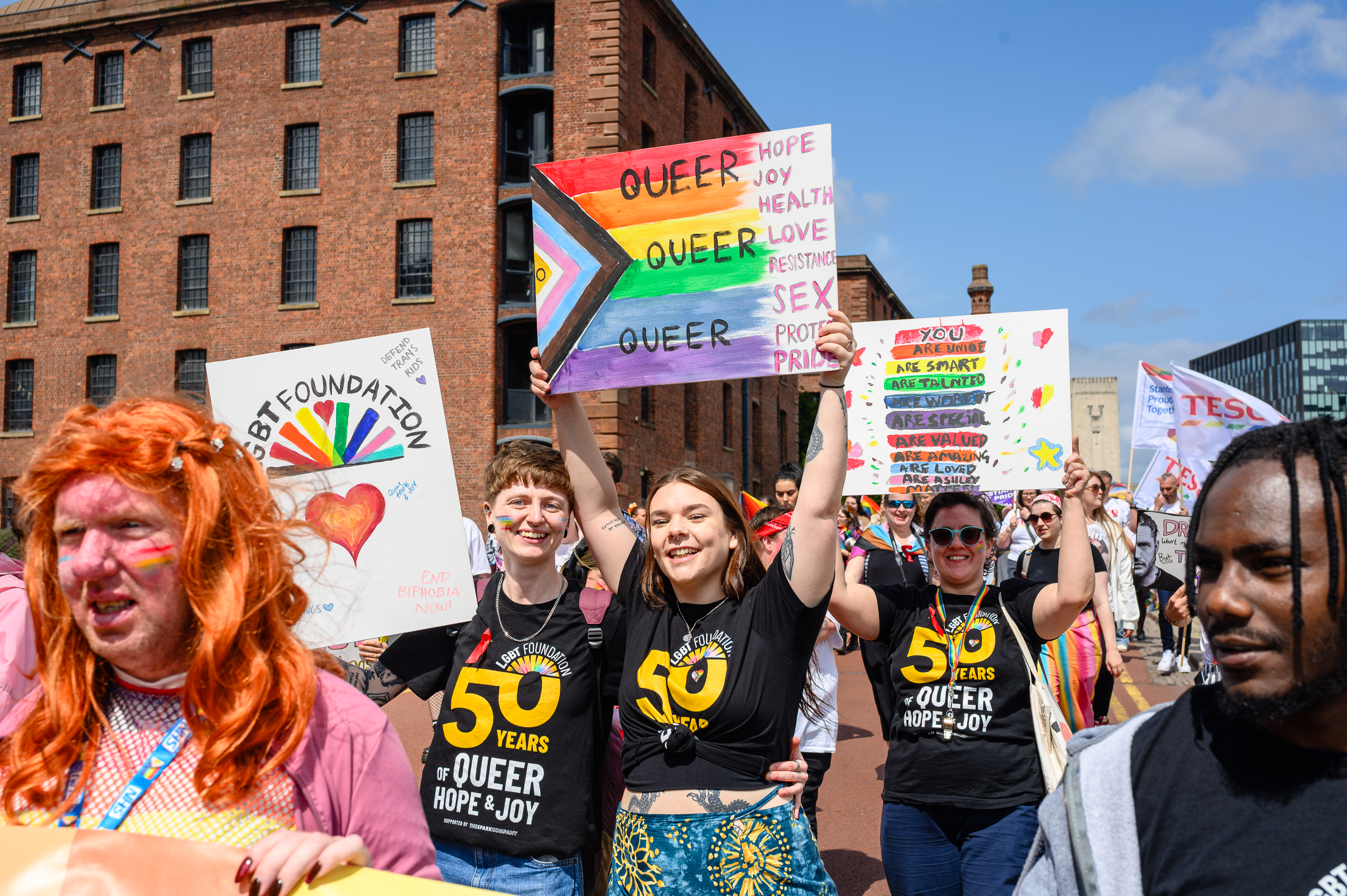 A group of LGBTQ+ advocates marching in the Liverpool Pride Parade. The focal people are wearing "50 Years of Queer Hope & Joy" LGBT Foundation t-shirts and holding placards in support of LGBTQ+ rights.