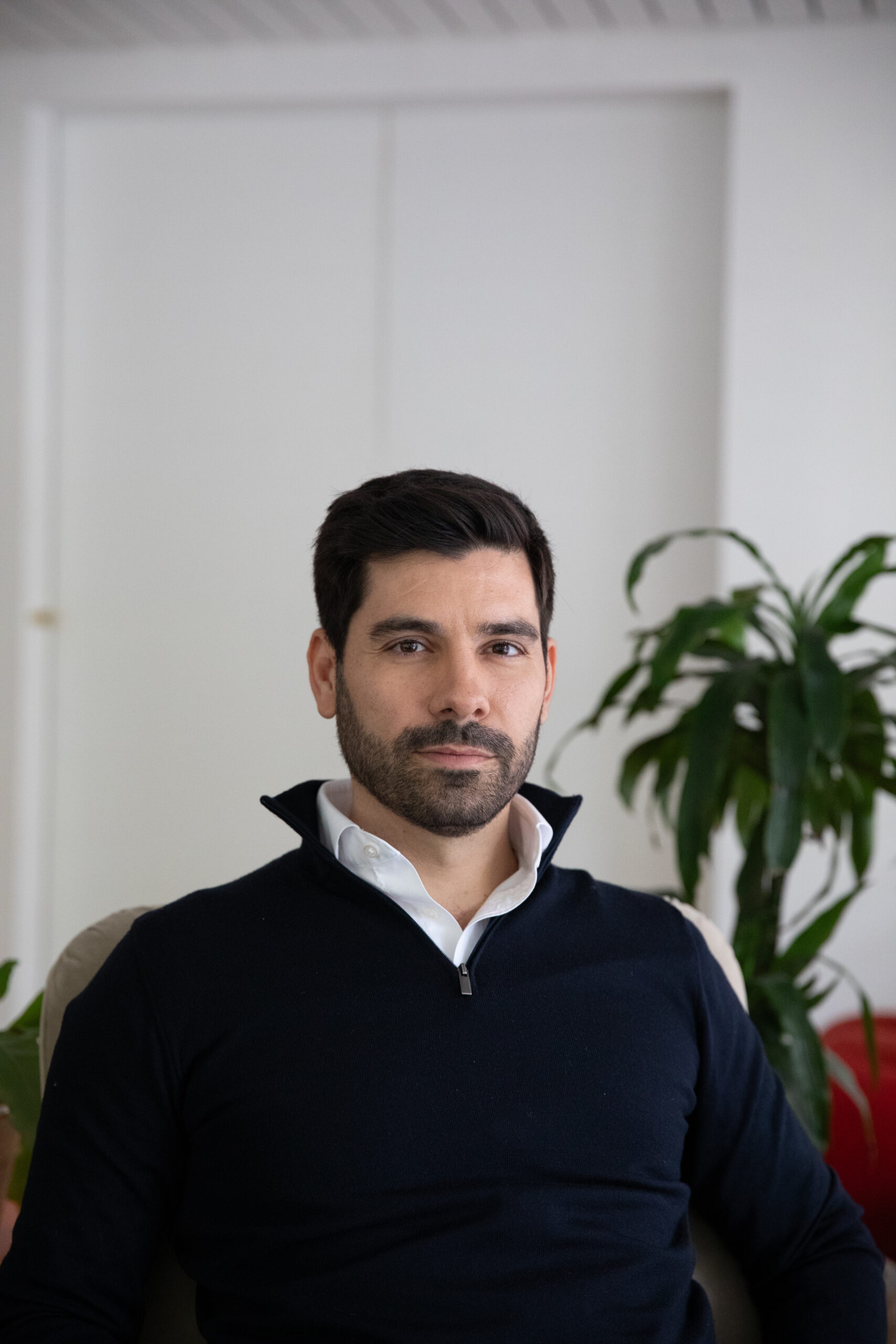 A portrait of our trustee Valerio. Valerio is European, masc-presenting with short dark hair, dark facial hair and is wearing a dark jumper with a white shirt with the collars raised. In the background is a white wall and a green potted plant.