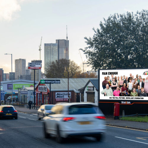 LGBT Foundation's Man Enough billboard, featuring a diverse group of LGBTQ+ men smiling against a white background, is visible as cars drive along a road into the distance.