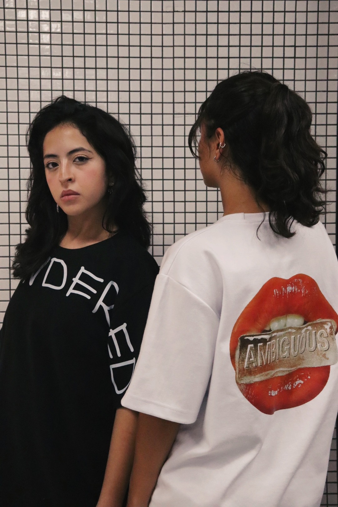 Two people against a checkered background wearing graphic tees. The person on the left is wearing a black tee with the slogan "UNGENDERED" on it in a white handwriting font and they are facing the camera. The person on the right is facing away from the camera and wearing a white t-shirt with a lips graphic on the back.