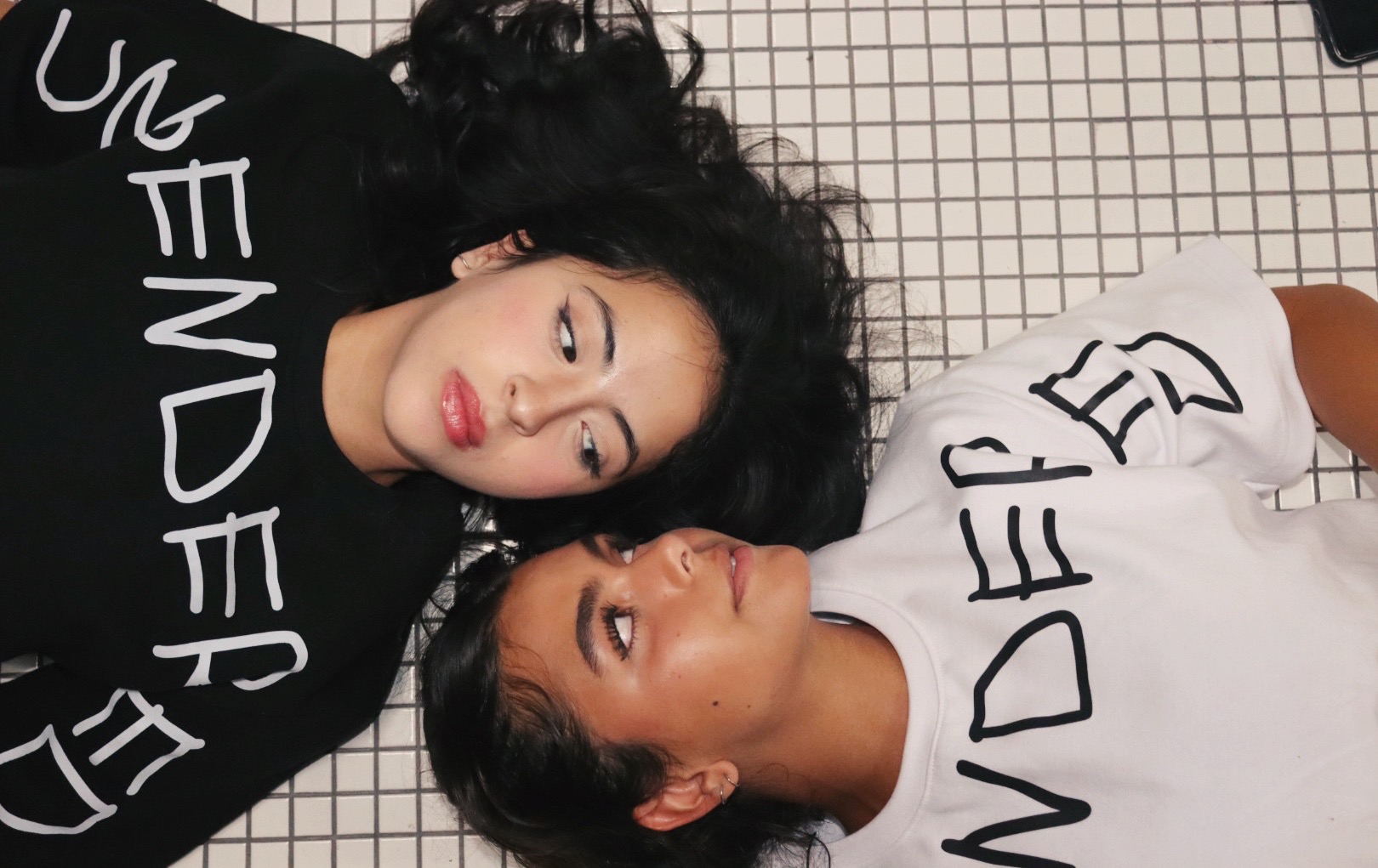 Two femme-presenting people lying down and looking at each other. The person on the top-left is wearing a black t-shirt with the slogan "UNGENDERED" on it, the person in the bottom-right is wearing a white version of the same t-shirt. They are lying against a checkered floor.