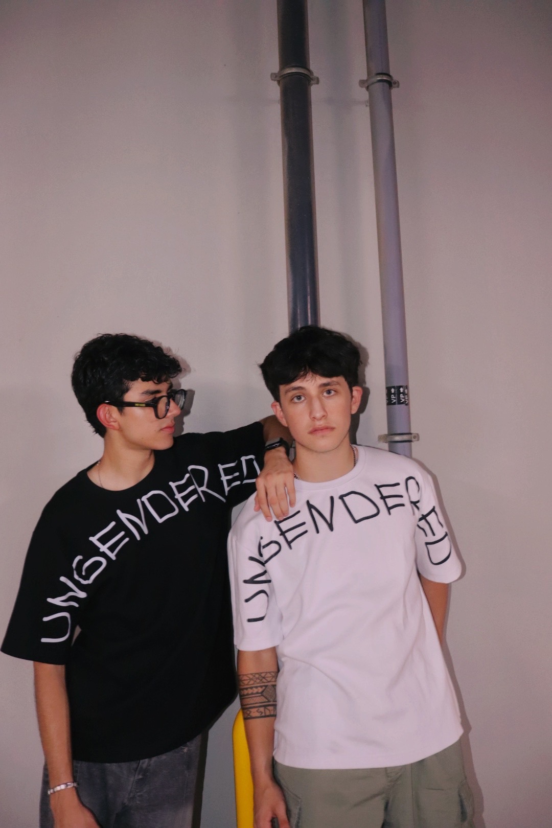 Two masc-presenting people wearing black and white graphic tees with the slogan "UNGENDERED" written across the upper chest area. The person on the left is wearing a black t-shirt and looking at the person on the right, who is facing the camera and wearing a white t-shirt.