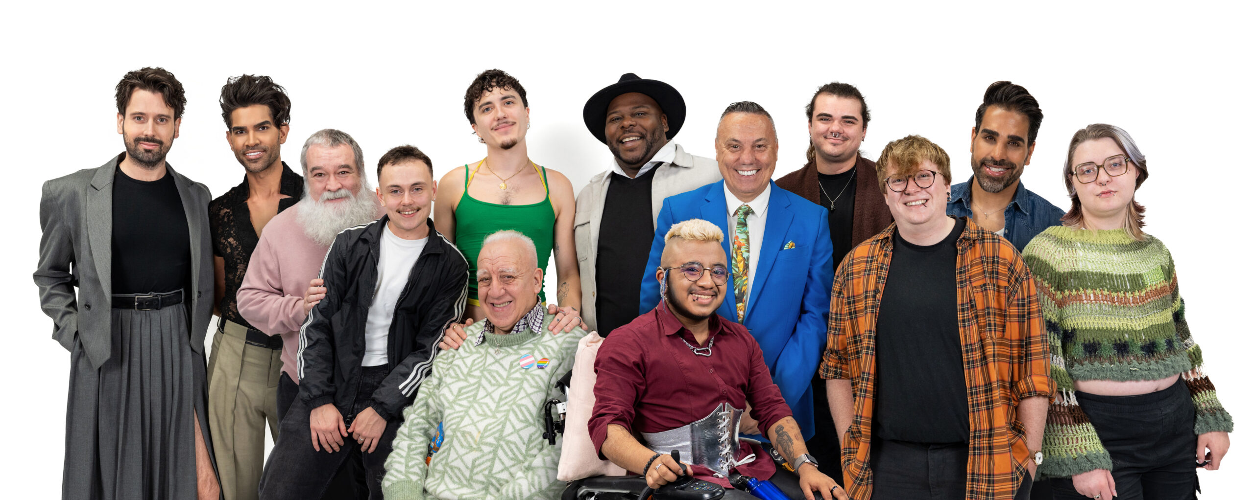 A diverse group of LGBTQ+ men looking at the camera and smiling in front of a white background. Several are standing and an older man and wheelchair user are centred in the foreground.