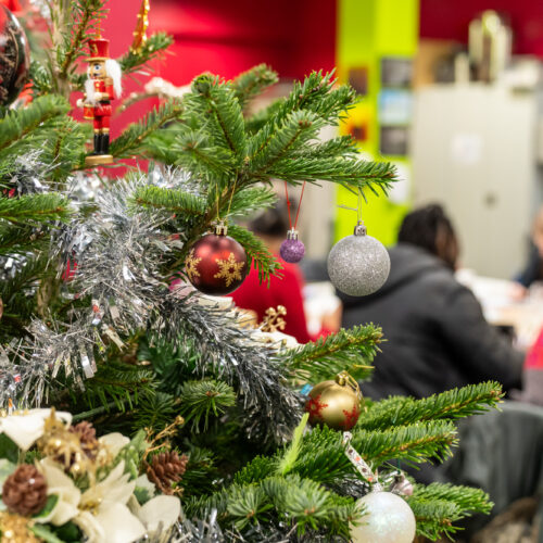 Christmas tree in the foreground with a group of people visible, out-of-focus and sitting around a table behind the tree.